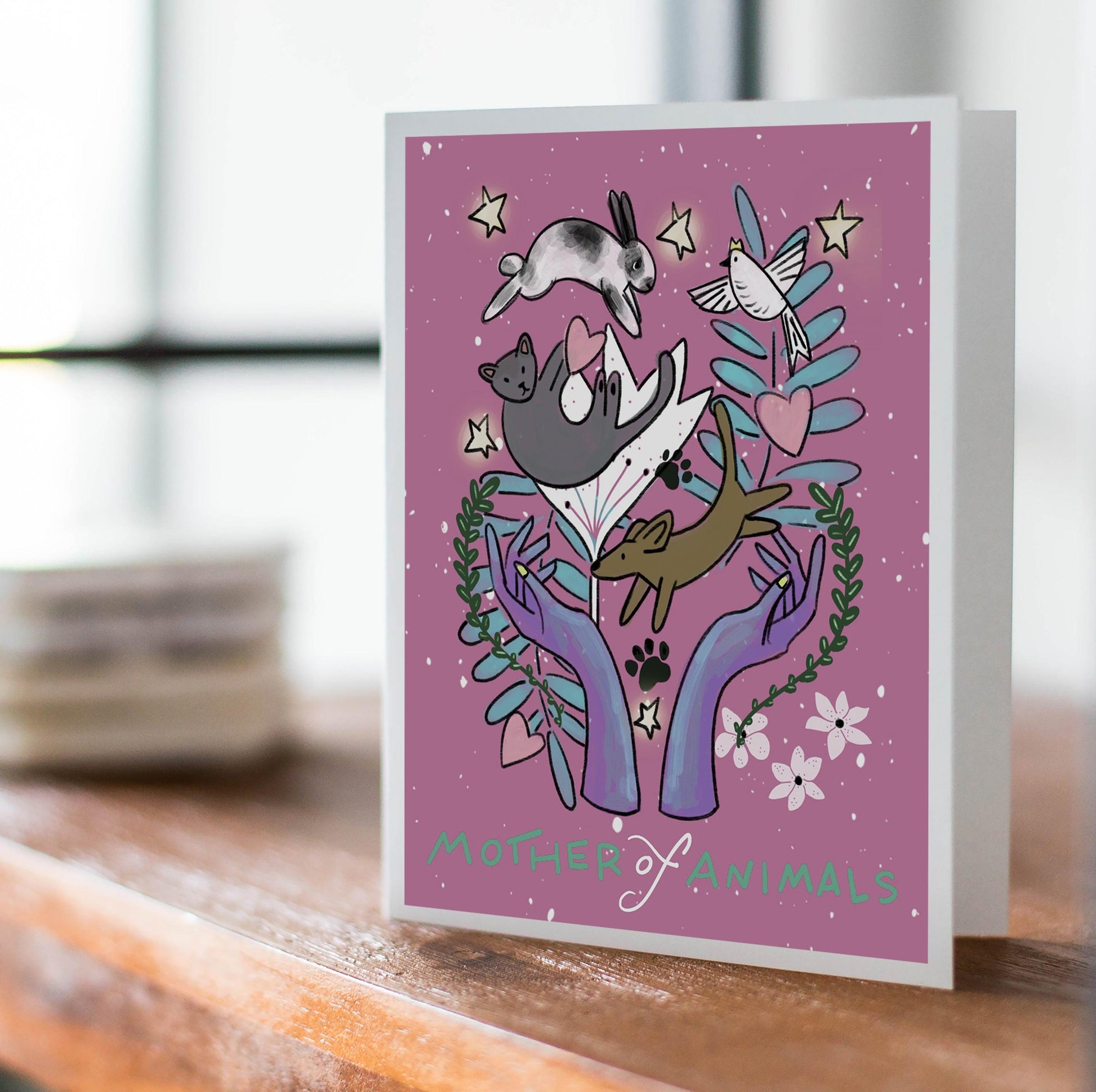 Pink card with two feminine hands holding whimsically drawn dog, cat, bunny, and bird with leaves and stars. Text below reads Mother of Animals